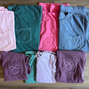 May include: A selection of colourful t-shirts in shades like pink, green, and blue. Each shirt has a pocket with a white embroidered Mickey Mouse design. The shirts are folded and arranged on a wooden surface.