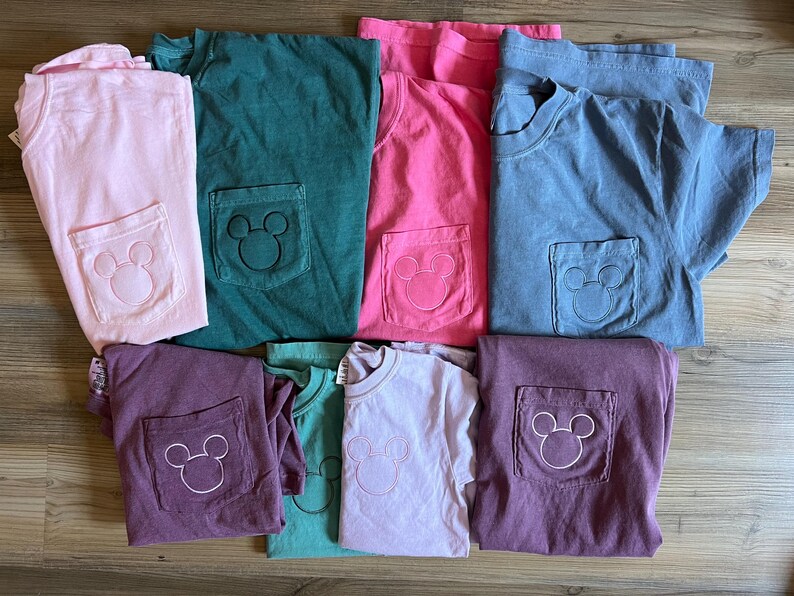 May include: A selection of colourful t-shirts in shades like pink, green, and blue. Each shirt has a pocket with a white embroidered Mickey Mouse design. The shirts are folded and arranged on a wooden surface.