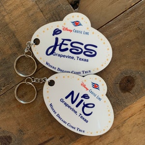 May include: Two white oval-shaped Disney Cruise Line name tags with gold stars and a blue ship logo. The top tag says "JESS Grapevine, Texas WHERE DREAMS COME TRUE". The bottom tag says "NIE Grapevine, Texas WHERE DREAMS COME TRUE".