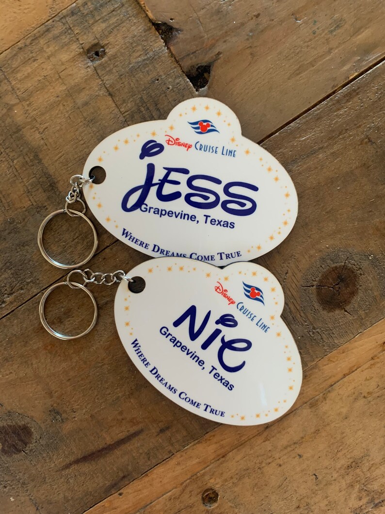 May include: Two white oval-shaped Disney Cruise Line name tags with gold stars and a blue ship logo. The top tag says "JESS Grapevine, Texas WHERE DREAMS COME TRUE". The bottom tag says "NIE Grapevine, Texas WHERE DREAMS COME TRUE".