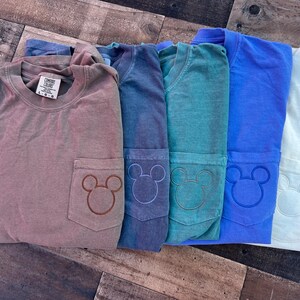 May include: Several Comfort Colors long-sleeve shirts in shades of brown, blue, teal, and white. Each shirt has a pocket with a Mickey Mouse design. The shirts are neatly folded and displayed on a wooden surface.