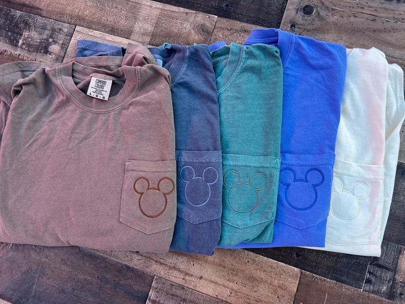 May include: Several Comfort Colors long-sleeve shirts in shades of brown, blue, teal, and white. Each shirt has a pocket with a Mickey Mouse design. The shirts are neatly folded and displayed on a wooden surface.