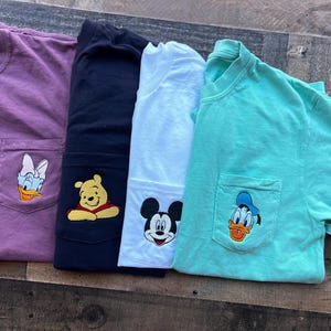 May include: Four short-sleeved t-shirts in purple, navy, white, and teal. Each shirt has a pocket with an embroidered cartoon character: Daisy Duck, Winnie the Pooh, Mickey Mouse, and Donald Duck. The shirts are folded on a wooden surface.