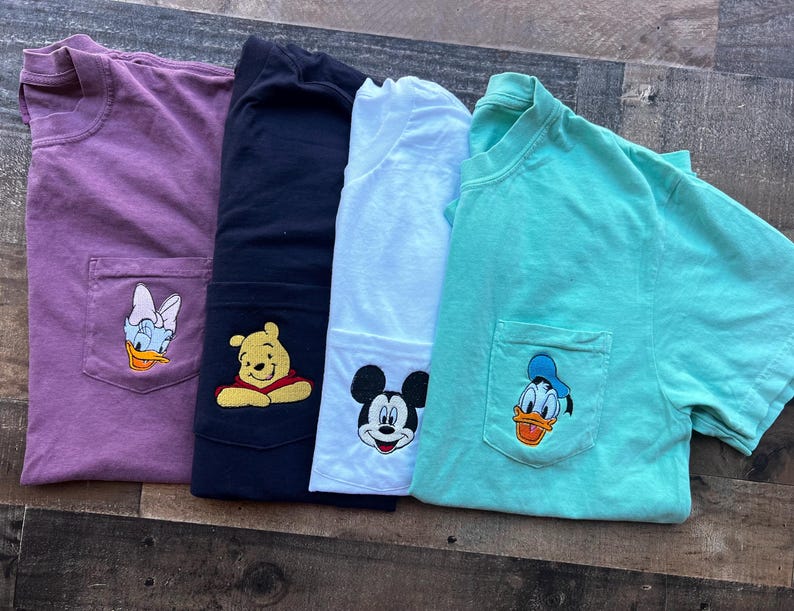 May include: Four short-sleeved t-shirts in purple, navy, white, and teal. Each shirt has a pocket with an embroidered cartoon character: Daisy Duck, Winnie the Pooh, Mickey Mouse, and Donald Duck. The shirts are folded on a wooden surface.
