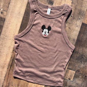 May include: A brown crop top with a classic Mickey Mouse embroidered design. The sleeveless top features a round neckline and a fitted silhouette. The Mickey Mouse design is in black, white, and red.