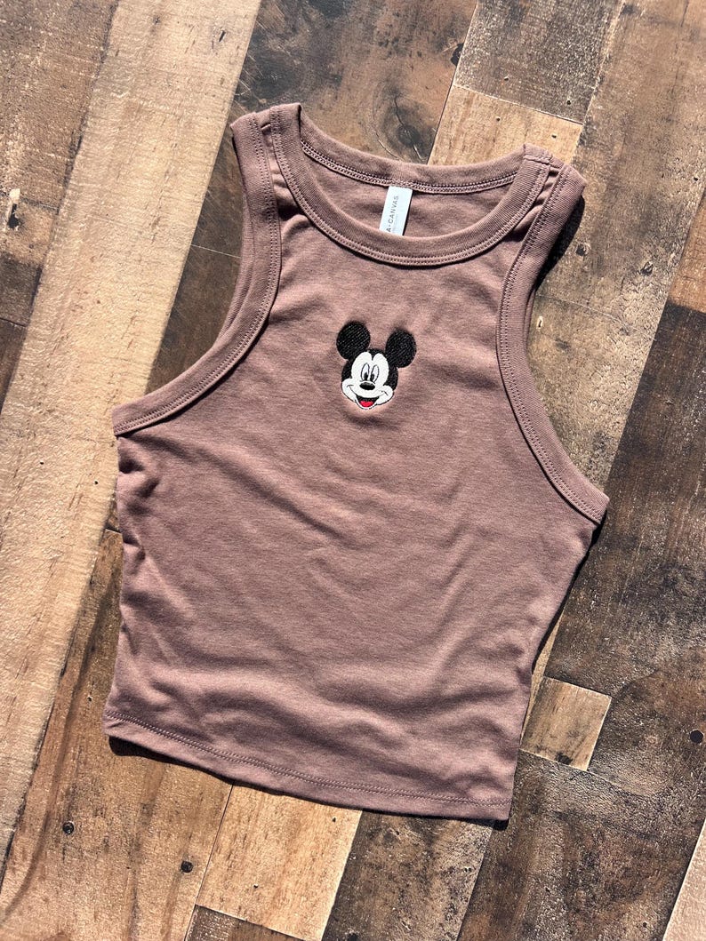 May include: A brown crop top with a classic Mickey Mouse embroidered design. The sleeveless top features a round neckline and a fitted silhouette. The Mickey Mouse design is in black, white, and red.