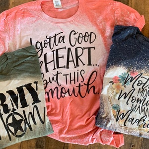 May include: Three bleached t-shirts with different designs. The first shirt is green with the text "Army Mom" in black. The second shirt is coral with the text "I gotta good heart... but this mouth." in black. The third shirt is navy blue with a floral design and the text "Mother Mom Mommy Mama Madre" in black.