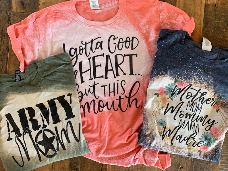 May include: Three bleached t-shirts with different designs. The first shirt is green with the text "Army Mom" in black. The second shirt is coral with the text "I gotta good heart... but this mouth." in black. The third shirt is navy blue with a floral design and the text "Mother Mom Mommy Mama Madre" in black.