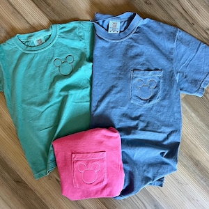 May include: Three short-sleeved t-shirts in teal, blue, and pink. Each shirt features a Mickey Mouse outline. The teal shirt has the design on the chest, while the blue and pink shirts have the design on the pocket.