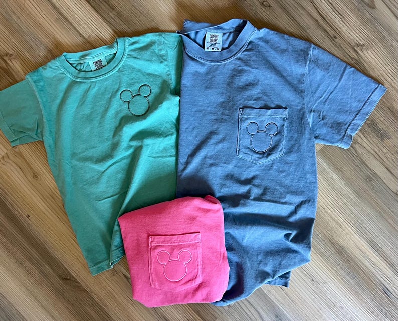 May include: Three short-sleeved t-shirts in teal, blue, and pink. Each shirt features a Mickey Mouse outline. The teal shirt has the design on the chest, while the blue and pink shirts have the design on the pocket.
