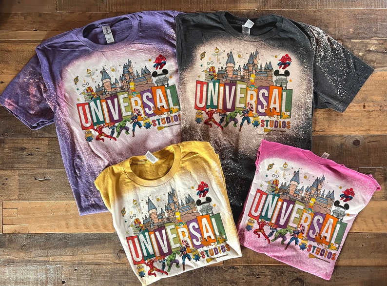 May include: Four bleached t-shirts featuring a graphic print of a castle, characters, and the text "Universal Studios". The shirts are in different colours: purple, black, yellow, and pink.