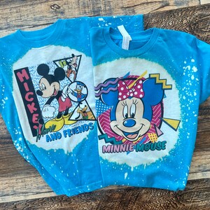 Puede incluir: Dos camisetas azules con dise&ntilde;os salpicados de lej&iacute;a. Una presenta a Mickey Mouse y el texto "Mickey Mouse and Friends". La otra presenta a Minnie Mouse y el texto "Minnie Mouse".