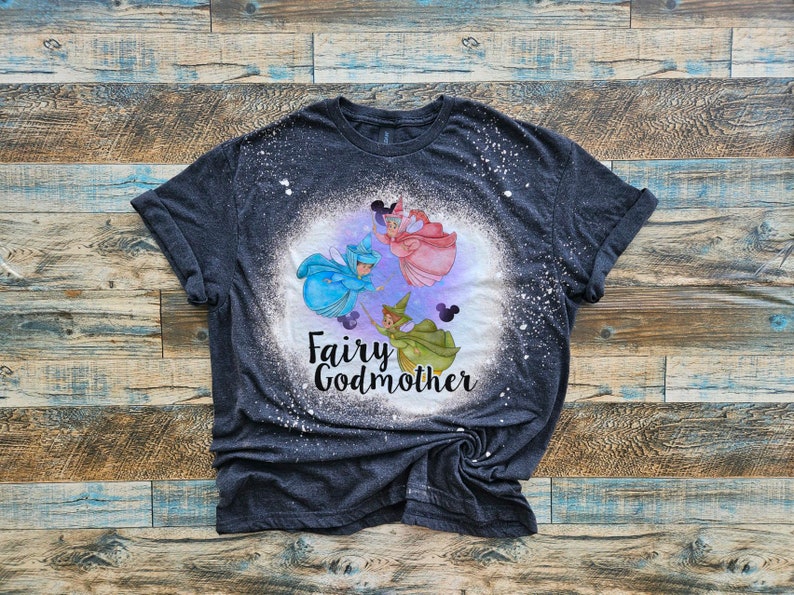 May include: A dark gray t-shirt with a bleach-splattered design featuring three fairies with wands and the text "Fairy Godmother".