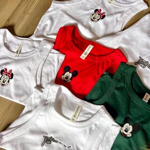 May include: A collection of white, red, and green tank tops and t-shirts. Some shirts feature embroidered cartoon character designs, including Minnie Mouse and Mickey Mouse. One white tank top has a gun design with the text "pew pew pew".