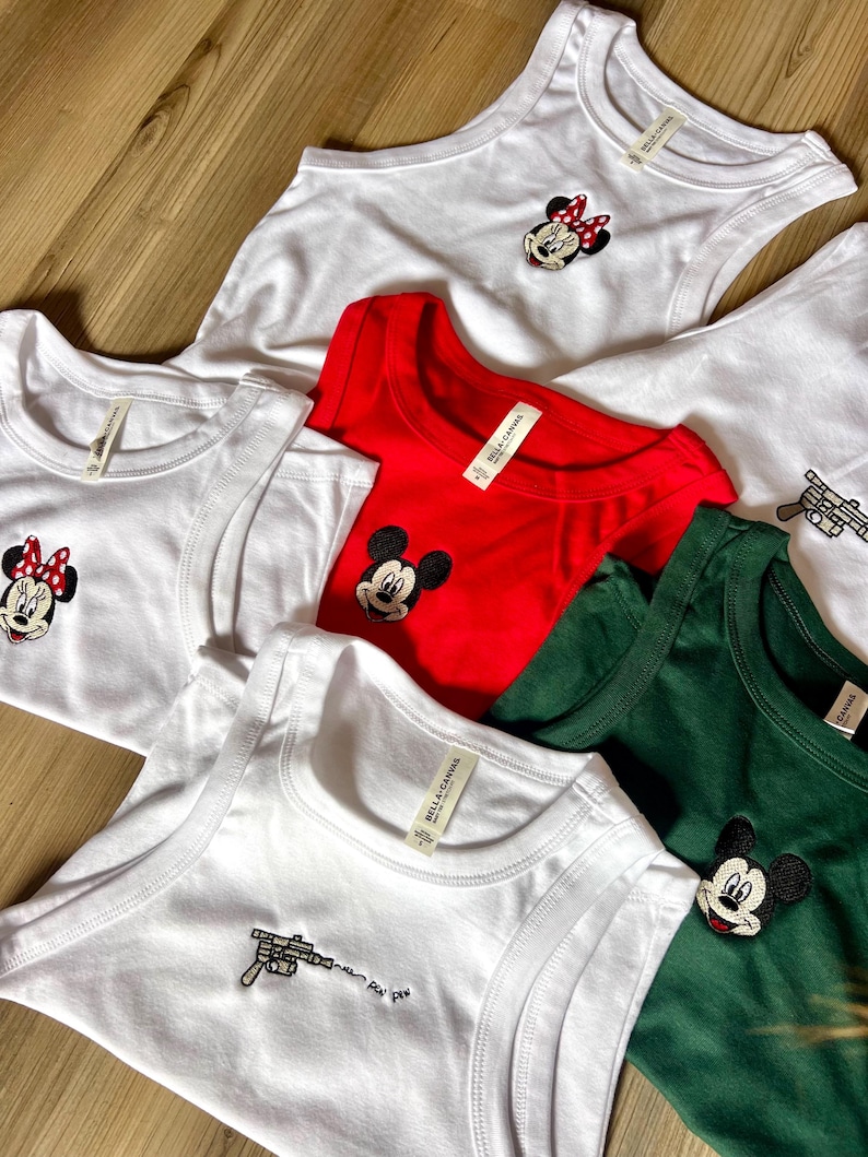 May include: A collection of white, red, and green tank tops and t-shirts. Some shirts feature embroidered cartoon character designs, including Minnie Mouse and Mickey Mouse. One white tank top has a gun design with the text "pew pew pew".