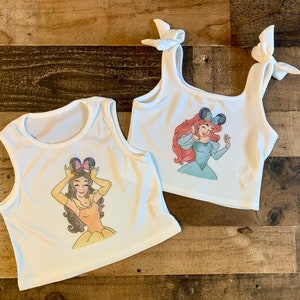K&ouml;nnte beinhalten: Zwei wei&szlig;e gerippte Tanktops. Ein Oberteil zeigt eine Grafik einer Figur mit braunen Haaren, die Mausohren und ein gelbes Kleid tr&auml;gt. Das andere Oberteil hat Schulterb&auml;nder zum Binden und eine Grafik einer Figur mit roten Haaren und Mausohren.
