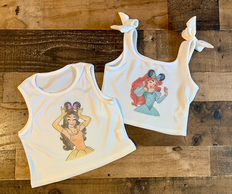 K&ouml;nnte beinhalten: Zwei wei&szlig;e gerippte Tanktops. Ein Oberteil zeigt eine Grafik einer Figur mit braunen Haaren, die Mausohren und ein gelbes Kleid tr&auml;gt. Das andere Oberteil hat Schulterb&auml;nder zum Binden und eine Grafik einer Figur mit roten Haaren und Mausohren.