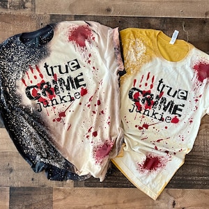Könnte beinhalten: Zwei weiße T-Shirts mit schwarzem Text, der "true crime junkie" lautet, und einem blutigen Handabdruck-Design. Ein Shirt hat ein schwarz-weißes Sprühdesign und das andere ein gelbes Sprühdesign.