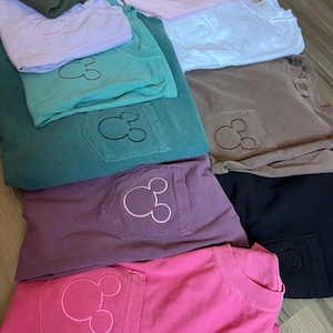 May include: An assortment of folded t-shirts in shades of pink, purple, green, and grey. Each shirt has a pocket with an embroidered Mickey Mouse design. The shirts are stacked on a light-coloured wooden surface.