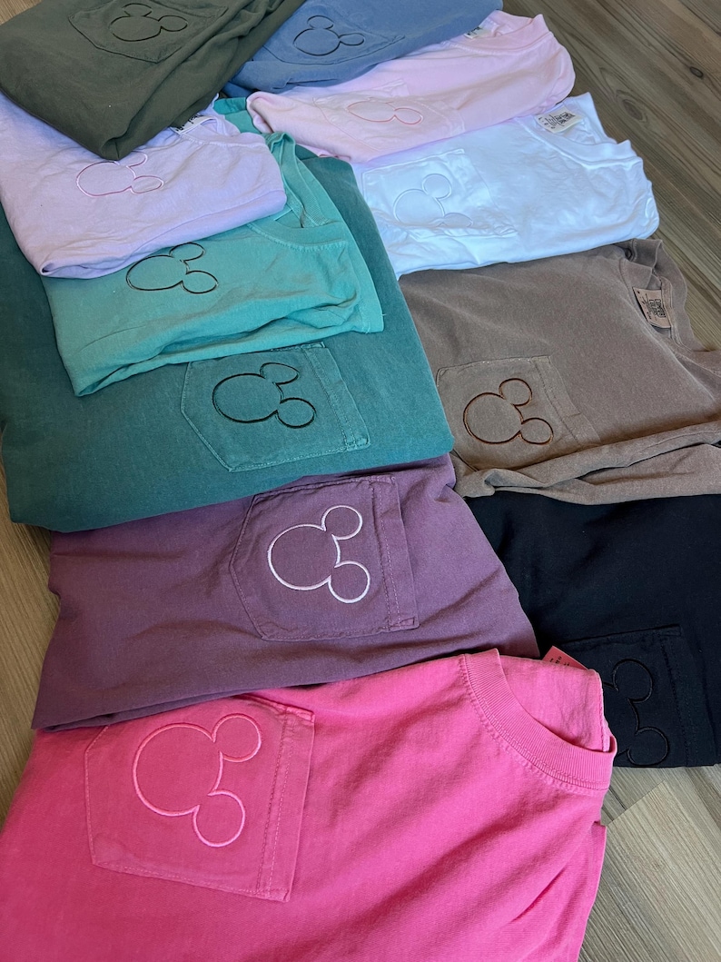 May include: An assortment of folded t-shirts in shades of pink, purple, green, and grey. Each shirt has a pocket with an embroidered Mickey Mouse design. The shirts are stacked on a light-coloured wooden surface.