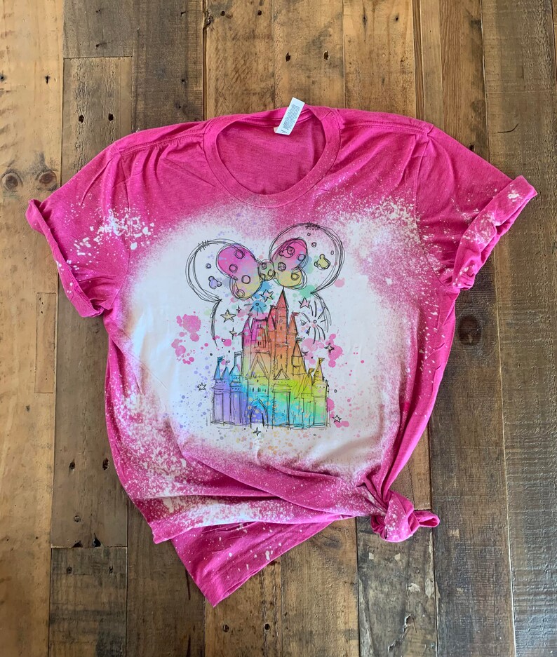 May include: Pink t-shirt with a bleach-splattered design featuring a rainbow-colored castle with Mickey Mouse ears and fireworks.