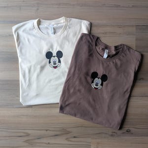 May include: Two t-shirts, one cream-colored and one brown, each featuring an embroidered Mickey Mouse face. The shirts are laid flat on a wooden surface, showcasing the classic cartoon character's iconic design.