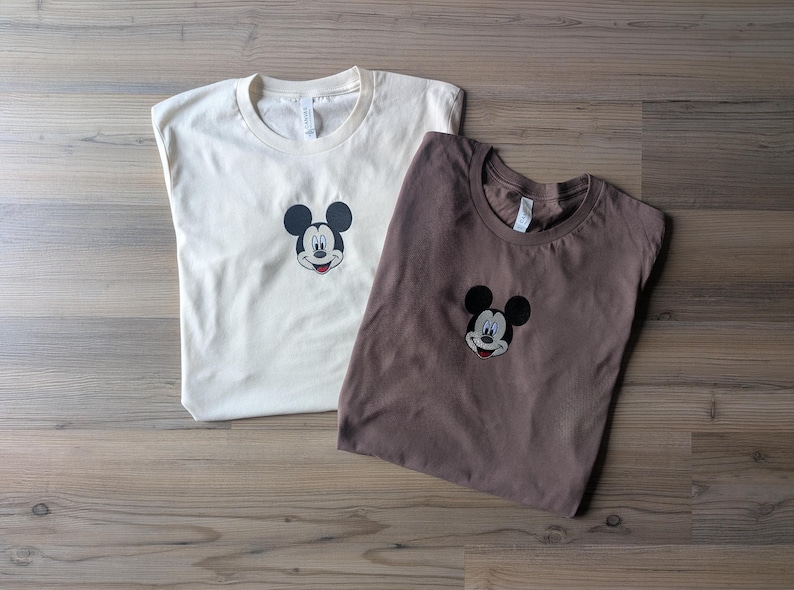 May include: Two t-shirts, one cream-colored and one brown, each featuring an embroidered Mickey Mouse face. The shirts are laid flat on a wooden surface, showcasing the classic cartoon character's iconic design.