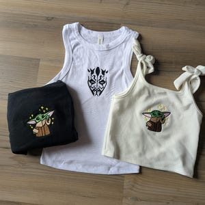 May include: A black t-shirt with an embroidered green alien character, a white tank top featuring a black graphic of a face with horns, and a cream-colored ribbed tank top with tie straps and an embroidered green alien character.