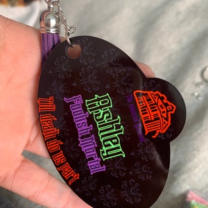 May include: A black oval-shaped keychain with a purple tassel. The keychain features a Haunted Mansion logo with the text "Disney's Haunted Mansion,  Foolish Mortal,  It's death do us part".