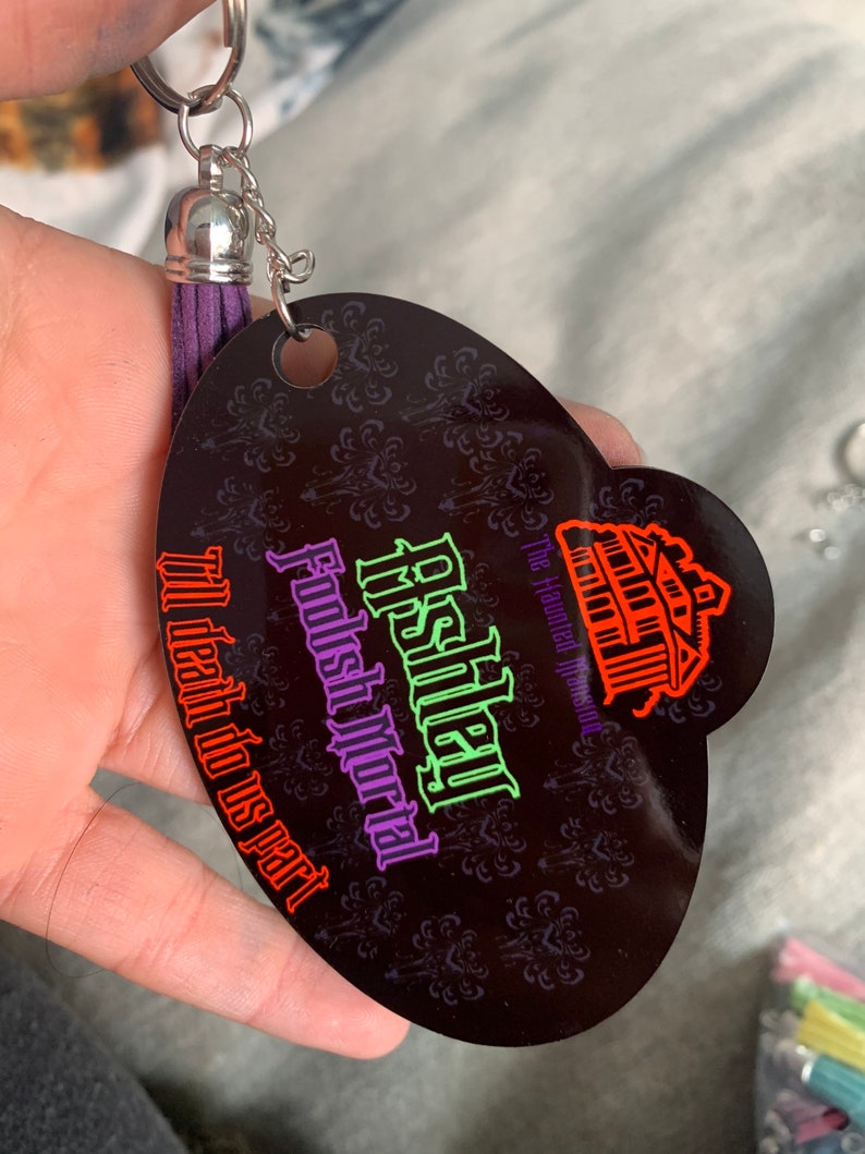 May include: A black oval-shaped keychain with a purple tassel. The keychain features a Haunted Mansion logo with the text "Disney's Haunted Mansion,  Foolish Mortal,  It's death do us part".
