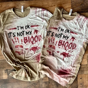 Op de afbeelding: Twee T-shirts met een verweerde, bespatte look. De shirts zijn licht khaki van kleur met rode spatten en de tekst "I'm OK It's Not My Blood" in zwart.