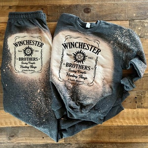 K&ouml;nnte beinhalten: Ein graues Sweatshirt- und Jogginghosen-Set mit einem Design aus Bleichmittelspritzern. Das Design zeigt eine schwarz-wei&szlig;e Grafik eines Etiketts mit der Aufschrift "Winchester Since 1983 Brothers Saving People Hunting Things A Family Business".