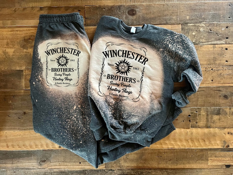 K&ouml;nnte beinhalten: Ein graues Sweatshirt- und Jogginghosen-Set mit einem Design aus Bleichmittelspritzern. Das Design zeigt eine schwarz-wei&szlig;e Grafik eines Etiketts mit der Aufschrift "Winchester Since 1983 Brothers Saving People Hunting Things A Family Business".