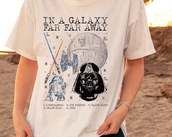 May include: White t-shirt with a graphic print of Star Wars characters and the text "IN A GALAXY FAR FAR AWAY". The characters are numbered and labeled: 1. Lightsabers, 2. Death Star, 3. Tie Fighter, 4. R2D2, 5. Darth Vader.