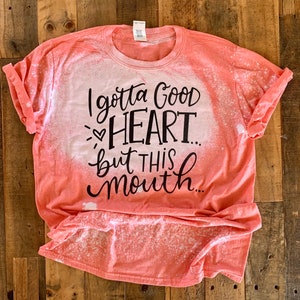 May include: A coral-colored t-shirt with a bleach-splattered design. The shirt has a black graphic that reads "I gotta good heart... but this mouth..."
