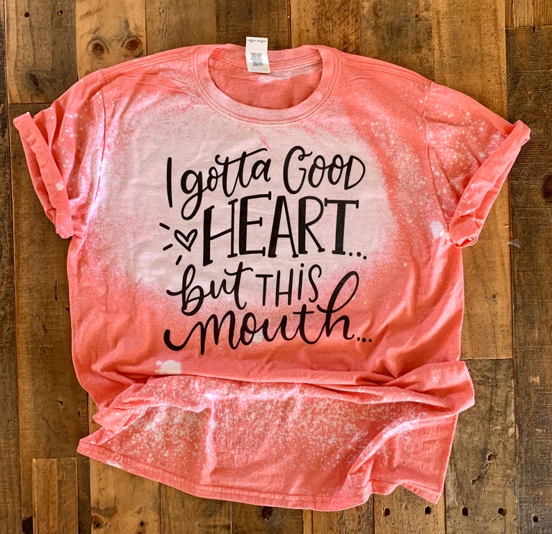 May include: A coral-colored t-shirt with a bleach-splattered design. The shirt has a black graphic that reads "I gotta good heart... but this mouth..."