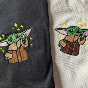 May include: Two shirts, one black and one white, each featuring an embroidered green creature with large ears wearing a brown robe. The character is depicted with one hand raised and is surrounded by small, yellow, four-pointed star-shaped accents.