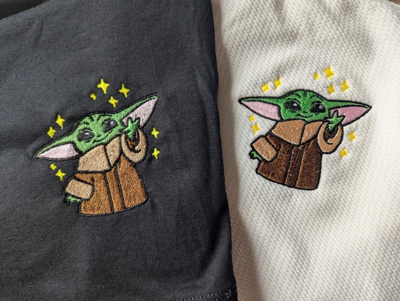 May include: Two shirts, one black and one white, each featuring an embroidered green creature with large ears wearing a brown robe. The character is depicted with one hand raised and is surrounded by small, yellow, four-pointed star-shaped accents.