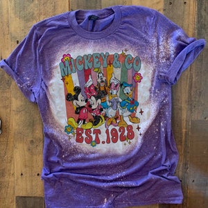 May include: Purple t-shirt with a bleached design featuring Mickey Mouse and friends. The design includes the text "Mickey & Co. Est. 1928".