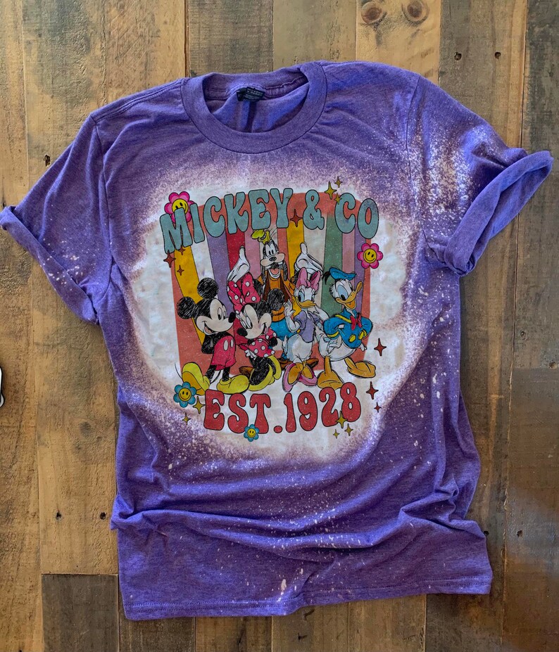 May include: Purple t-shirt with a bleached design featuring Mickey Mouse and friends. The design includes the text "Mickey & Co. Est. 1928".