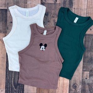 May include: Three sleeveless tank tops in cream, brown, and dark green. The brown tank top features an embroidered Mickey Mouse design. The tops are made of a soft, ribbed material, perfect for casual wear.