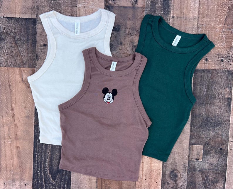 May include: Three sleeveless tank tops in cream, brown, and dark green. The brown tank top features an embroidered Mickey Mouse design. The tops are made of a soft, ribbed material, perfect for casual wear.