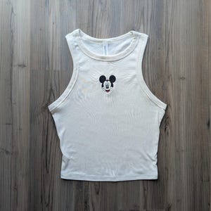 May include: Cream-colored tank top featuring a small embroidered Mickey Mouse face on the chest. The sleeveless top has a rounded neckline and is made of a soft, ribbed fabric. The tank top is a casual style.