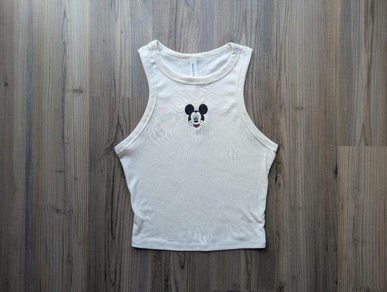 May include: Cream-colored tank top featuring a small embroidered Mickey Mouse face on the chest. The sleeveless top has a rounded neckline and is made of a soft, ribbed fabric. The tank top is a casual style.