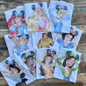 K&ouml;nnte beinhalten: Eine Sammlung wei&szlig;er T-Shirts mit farbenfrohen grafischen Illustrationen verschiedener animierter Prinzessinnen, die Mausohr-Haarreifen tragen. Jedes T-Shirt zeigt eine andere Figur mit individueller Haarfarbe, Kleidung und Gesichtsausdruck.