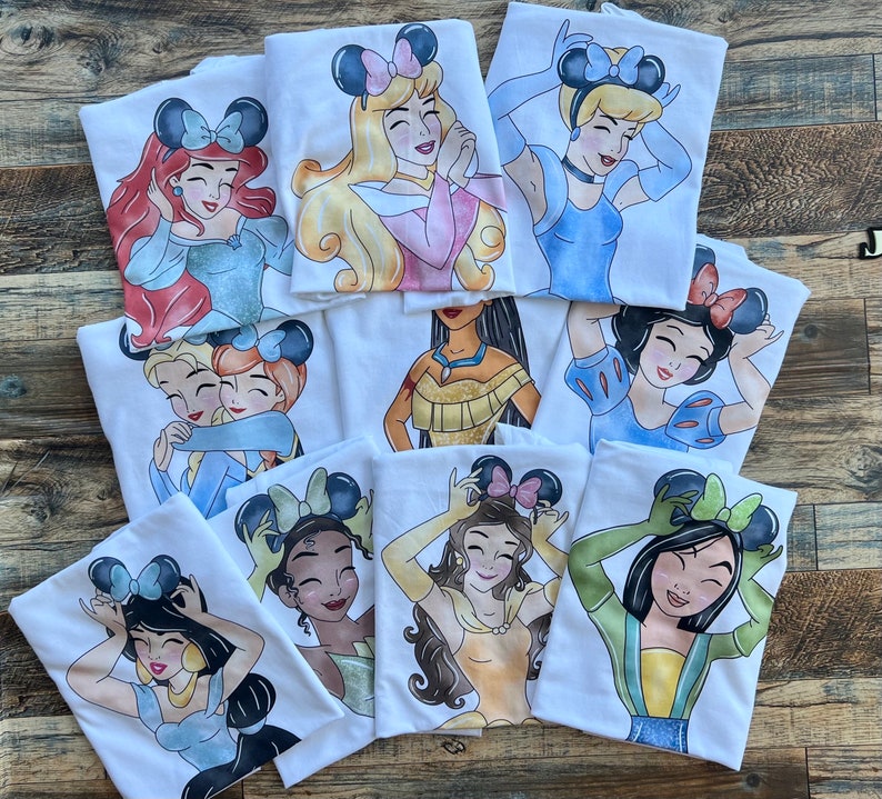 K&ouml;nnte beinhalten: Eine Sammlung wei&szlig;er T-Shirts mit farbenfrohen grafischen Illustrationen verschiedener animierter Prinzessinnen, die Mausohr-Haarreifen tragen. Jedes T-Shirt zeigt eine andere Figur mit individueller Haarfarbe, Kleidung und Gesichtsausdruck.