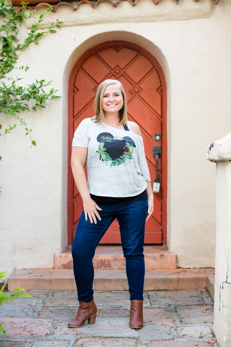 May include: A woman wearing a white t-shirt with a black and green Mickey Mouse design, blue jeans, and brown ankle boots. She is standing in front of a red door.
