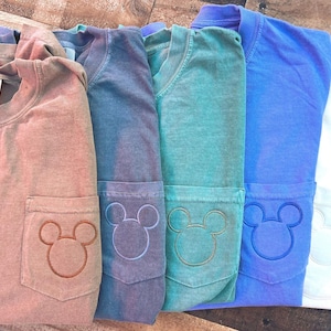 May include: Several Comfort Colors long-sleeve shirts in shades of brown, blue, and green. Each shirt has a pocket with an embroidered Mickey Mouse design. The shirts are neatly folded and presented on a wooden surface.