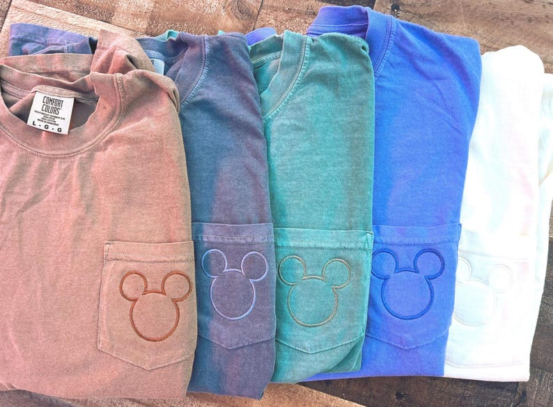 May include: Several Comfort Colors long-sleeve shirts in shades of brown, blue, and green. Each shirt has a pocket with an embroidered Mickey Mouse design. The shirts are neatly folded and presented on a wooden surface.