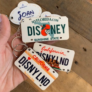 May include: Three wooden keychains shaped like license plates. The top keychain is white with blue text that reads "MYFLORIDA.COM DISNEY SUNSHINE STATE". The middle keychain is white with blue text that reads "California DISNYLND". The bottom keychain is white with orange text that reads "CALIFORNIA DISNY LND The Golden State".